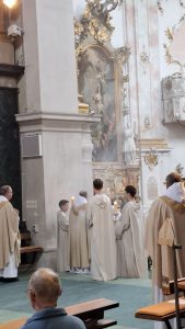 Innenansicht einer Kirche während eines Gottesdienstes. Mehrere Geistliche und Messdiener in hellen, beigen Gewändern stehen in der Bildmitte vor einem reich verzierten Altar, auf dem Kerzen brennen und Weihrauch aufsteigt. Der Altarbereich ist mit einem großen Gemälde und goldenen Ornamenten geschmückt. Links im Bild steht ein weiterer Geistlicher. Im Vordergrund sitzt ein Mann mit Brille und Glatze, von hinten gesehen. Der Boden ist mit grünem Teppich ausgelegt, und hohe Säulen sowie kunstvolle Wandverzierungen sind im Hintergrund sichtbar.