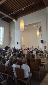 Eine große Innenansicht eines Saals oder einer Kirche während einer musikalischen Aufführung. Im Zentrum stehen junge Musiker mit Blasinstrumenten und ein Dirigent auf einer Bühne. Im Vordergrund sitzt ein Publikum, hauptsächlich Jugendliche, mit dem Rücken zur Kamera. Der Raum hat eine hohe Holzdecke mit sichtbaren Balken, große Hängelampen, weiße Wände, ein hohes Fenster und ein großes Wandgemälde auf der linken Seite.