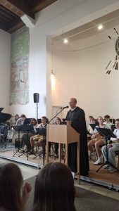 Ein Sprecher in schwarzer Robe steht an einem hölzernen Rednerpult auf einer Bühne und gestikuliert mit seiner rechten Hand. Hinter ihm sitzt eine Gruppe Jugendlicher mit Musikinstrumenten wie Saxophonen, einem Waldhorn und einem Schlagzeug um Notenständer herum. Links im Hintergrund ist ein großes, verwittertes Wandgemälde in Grün- und Brauntönen zu sehen. Der hohe Raum hat helle Wände, eine dunkle Holzbalkendecke links und moderne Lichtschienen mit Strahlern rechts. Im Vordergrund sind die Hinterköpfe von zwei Zuschauern zu erkennen, die auf einem Teppichboden sitzen.