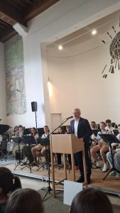 Ein Mann mit grauem Haar und Brille in einem dunklen Anzug spricht von einem hölzernen Pult aus in ein Mikrofon. Hinter ihm auf einer erhöhten Bühne ist eine Gruppe junger Musiker mit Notenständern, Saxophonen und einem Schlagzeug platziert. Der hohe Raum hat eine dunkle Holzdecke und helle Wände. Links an der Wand befindet sich eine große, verblasste Wandmalerei, rechts eine moderne Lichtinstallation mit Strahlern. Im unteren Vordergrund sind die Köpfe von Zuschauern sichtbar.