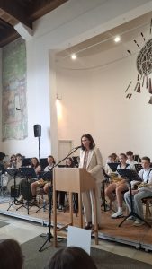 Eine Frau in einem hellen Anzug spricht an einem hölzernen Rednerpult in einem großen Raum. Hinter ihr sitzt eine Gruppe Jugendlicher mit Musikinstrumenten wie Saxophonen und einem Schlagzeug auf einer erhöhten Bühne. Der Raum hat hohe, helle Wände, wobei links ein großes, verblasstes Wandgemälde und rechts oben ein abstraktes Kunstwerk sichtbar sind. An der Decke sind Holzbalken und eine Lichtleiste mit Strahlern angebracht. Der Boden im Vordergrund ist teppichbelegt, die Bühne ist aus Holz.