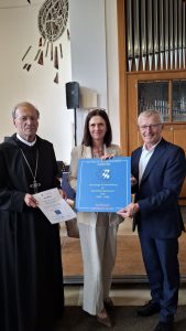 Drei Personen stehen in einem Innenraum mit hoher Decke und Holzvertäfelung. Links steht ein Mann in schwarzer Mönchskutte mit Brustkreuz, der ein weißes Zertifikat mit der Aufschrift „Zertifikat“ und „Benediktinergymnasium Ettal“ hält. In der Mitte steht eine Frau in beiger Kleidung, rechts ein Mann im dunklen Anzug. Beide halten gemeinsam ein blaues Schild hoch. Auf dem Schild steht „KATHOLISCHES SCHULWERK - IN BAYERN“, darunter ein Logo mit Kreuz und bayerischen Rauten, sowie „Nachhaltige Schulentwicklung am Benediktinergymnasium Ettal (2024 - 2028)“ und „Zertifizierte Katholische Schule“. Im Hintergrund sind eine große Orgel mit Pfeifen und ein modernes Wandkunstwerk zu sehen.