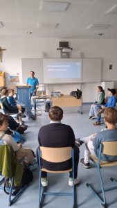 Ein Mann in einem blauen Poloshirt steht in einem Klassenzimmer und präsentiert vor einer Gruppe von Schülern. Auf einer großen weißen Tafel hinter ihm wird ein Beamer-Bild mit dem Titel "Schülermedientag 2025", dem Untertitel "Medien und Formate" und dem Logo "BR" (Bayerischer Rundfunk) projiziert. Die Schülerinnen und Schüler sitzen auf blauen Stühlen in einem Halbkreis und blicken zum Redner. Der Raum hat einen hellblauen Boden, weiße Wände und eine weiße Rasterdecke. Links an der Wand hängt ein Holzkreuz. Auf einem Tisch vor dem Redner stehen ein Laptop, ein Beamer und weitere technische Geräte.