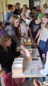 Eine Gruppe junger Menschen füllt an einem mit Zeitungen bedeckten Tisch Pommes Frites aus einer Metallschüssel in mehrere Pappbehälter. Zwei Frauen stehen am Tisch, eine davon trägt weiße Handschuhe und legt Pommes in die Verpackungen. Um den Tisch herum stehen weitere Jugendliche, darunter ein Mädchen in einem Dirndl und ein Junge mit grüner Kappe, die beobachten oder warten. Im Hintergrund ist eine große Landkarte an der Wand zu sehen, was auf ein Klassenzimmer oder einen Gemeinschaftsraum hindeutet.