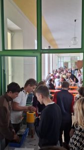 Eine belebte Szene in einer großen Halle oder Cafeteria, die durch grüne Fenster- und Türrahmen hindurch betrachtet wird. Im Vordergrund stehen mehrere Personen an einem Tisch mit Behältern und Utensilien, möglicherweise bei der Essensausgabe. Im Hintergrund sitzen und stehen zahlreiche Menschen an langen orangefarbenen Tischen in einem hellen Raum mit weißen Säulen. Die Decke ist weiß mit gelben Diagonalstreifen und einer großen, kugelförmigen Lampe.