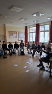 Ein Klassenzimmer, in dem eine Gruppe von etwa zehn jungen Männern in einem Halbkreis auf Stühlen sitzt. Ein erwachsener Mann sitzt ihnen gegenüber und gestikuliert mit Papieren in der Hand. Auf dem hellbraunen Fußboden liegen mehrere weiße Zettel. Im Hintergrund sind hellgelbe Wände mit zwei großen Pinnwänden zu sehen. Mehrere große Fenster mit roten Vorhängen lassen Tageslicht herein und bieten den Blick auf ein Gebäude außerhalb. Die weiße Decke ist mit Akustikplatten und quadratischen Deckenleuchten ausgestattet.