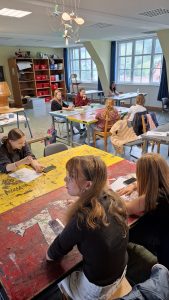 Innenaufnahme eines hellen Klassenzimmers oder Kunstraums, in dem mehrere junge Frauen an farbigen, bemalten Tischen sitzen. Im Vordergrund malt eine junge Frau ein Muster auf ihren Unterarm, daneben sitzt eine andere, die zuschaut. Im Mittelgrund sind weitere Schülerinnen an ihren Tischen bei verschiedenen Aktivitäten zu sehen. Im Hintergrund bieten große Fenster einen Blick auf eine grüne Landschaft mit Hügeln und Gebäuden. Links steht ein Holzregal mit roten Aufbewahrungsboxen. Von der Decke hängen kunstvolle Mobiles.