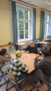 Fünf Personen, darunter ein älterer Mann und vier jüngere Männer, sitzen um einen quadratischen Holztisch und spielen ein Kartenspiel. Spielkarten und farbige Chips sind auf der Tischplatte ausgebreitet. Der ältere Mann hält Karten in den Händen und blickt auf das Spiel. Der Raum ähnelt einem Klassenzimmer mit großen, mehrteiligen Fenstern und blauen Vorhängen im Hintergrund. Durch die Fenster ist grünes Laubwerk sichtbar. Weitere Tische und Stühle sind im Raum verteilt.