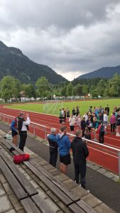 Außenaufnahme eines Sportplatzes mit roter Laufbahn und grünem Rasenfeld. Im Hintergrund erheben sich bewaldete Berge unter einem bewölkten Himmel. Auf der Laufbahn und dem Rasenfeld steht eine große Gruppe von Kindern und Jugendlichen, einige im Sportdress. Einige Fußballtore sind auf dem Rasenfeld sichtbar. Im Vordergrund stehen mehrere Erwachsene hinter einem Metallgeländer, das die Tribüne von der Laufbahn trennt. Ein Mann mit weißem Hemd und Weste gestikuliert in Richtung der Gruppe. Im unmittelbaren Vordergrund sind Holzbänke einer Tribüne zu sehen, auf einer liegt eine rote Tasche.