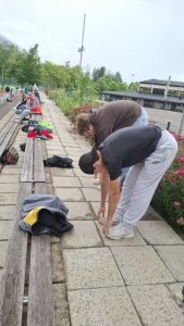 Zwei junge Männer beugen sich auf einem nassen Pflasterweg nach vorne. Der eine trägt ein schwarzes T-Shirt und eine graue Jogginghose, der andere einen braunen Kapuzenpullover und Jeans. Hinter ihnen verläuft eine lange Holzbank, auf der mehrere Personen sitzen und Taschen liegen. Neben dem Weg befindet sich ein Blumenbeet mit rosa Blüten. Im Hintergrund sind grüne Bäume, Berge, eine Sportbahn und Gebäude sichtbar.