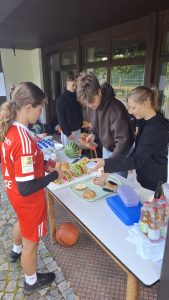 Vier Jugendliche bereiten draußen an einem langen weißen Tisch Snacks zu. Eine junge Frau in einem roten Sporttrikot hält ein Brötchen in der Hand. Auf dem Tisch liegen aufgeschnittene Wassermelone, weitere Brötchen, Wasser- und Saftflaschen. Zwei weitere Jugendliche schneiden Wassermelone und arrangieren Brötchen auf einem Tablett. Ein Basketball liegt auf dem gepflasterten Boden neben dem Tisch. Im Hintergrund sind Fenstertüren und Bäume zu sehen.