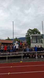 Eine Gruppe junger Menschen steht auf den Tribünenstufen einer Sportanlage und blickt in die Kamera. Der Himmel ist bedeckt. Links und rechts stehen hohe, schlanke Masten. Im Hintergrund befinden sich ein Holzhaus mit rotem Ziegeldach, ein kleines, modernes weißes Gebäude mit großen Fenstern, sowie grüne Bäume und bewaldete Berge. Im Vordergrund ist eine rote Laufbahn mit weißen Linien und gelben Markierungen zu sehen, durch ein Metallgeländer von den Personen getrennt.