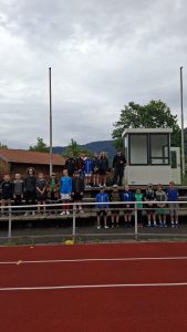 Eine Gruppe von etwa 20 Kindern und Jugendlichen steht in zwei Reihen auf einer Tribüne einer Sportanlage. Sie tragen unterschiedliche Sport- und Freizeitkleidung. Im Vordergrund ist eine rote Laufbahn mit weißen Linien zu sehen. Im Hintergrund erheben sich grüne, bewaldete Berge unter einem grauen, bewölkten Himmel. Links ist ein Gebäude mit einem braun-roten Dach sichtbar, und rechts steht eine kleine, weiße Kabine. Zwei hohe, schlanke Masten ragen in den Himmel.