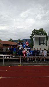 Ein Gruppenbild von etwa 22 jungen Männern, die auf den Stufen einer Tribüne auf einer Sportanlage stehen. Im Vordergrund ist eine rote Kunststofflaufbahn mit weißen Markierungen sichtbar. Rechts von der Gruppe steht eine weiße Kabine mit Fenstern, und links ist ein Gebäude mit rotem Dach zu sehen. Im Hintergrund erstreckt sich eine bewaldete Hügellandschaft unter einem bewölkten Himmel. Hohe Masten ragen links und rechts in den Himmel.
