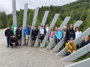 Eine Gruppe von etwa zwanzig Personen verschiedenen Alters steht auf einem Schotterweg vor einer Reihe von schrägen, grauen Metallstreben. Im Hintergrund erstreckt sich ein dichter Nadelwald unter einem bewölkten Himmel. Links neben der Gruppe steht ein grünes Informationsschild.