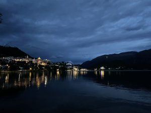 Eine Abenddämmerungsszene zeigt eine beleuchtete Stadt am linken Ufer eines dunklen Sees. Die warmen Lichter der Stadt spiegeln sich deutlich auf der ruhigen Wasseroberfläche. Darüber liegt ein dunkelblauer, stark bewölkter Himmel, und im Hintergrund erheben sich dunkle Bergketten.