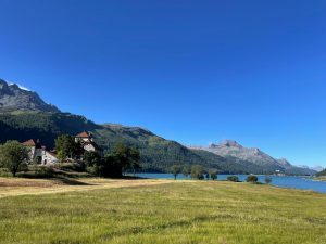 Ein weites Landschaftsbild zeigt im Vordergrund ein grünes Feld. Links steht ein großes Steingebäude mit roten Dächern, umgeben von Bäumen. Dahinter liegt ein leuchtend blauer See, an dessen Ufer vereinzelte Bäume zu sehen sind. Im Hintergrund erheben sich bewaldete Berge unter einem klaren, blauen Himmel.