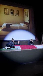Eine Theaterbühne mit einer großflächigen Projektion eines schlichten japanischen Schlafzimmers im Hintergrund, komplett mit einem Bett auf Tatami-Matten und gerahmten Bildern (u.a. Berg Fuji, Kirschblüte, Kranich) an den Wänden. Im Vordergrund knien auf der dunklen Bühne drei Personen in schwarzen Kostümen mit Ohrenbändern. Sie sind um zwei bettähnliche Objekte herum arrangiert, von denen eines mit einer roten Decke bedeckt ist. Ein großer, runder Scheinwerfer beleuchtet die Personen und den unteren Teil der Projektion.