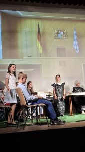 Eine Gruppe von Kindern inszeniert eine Szene auf einer Bühne mit grünem Kunstrasen. Im Hintergrund ist eine große Projektion eines Gerichtssaals oder offiziellen Raumes zu sehen. Diese Projektion zeigt die deutsche Flagge (schwarz-rot-gold), das bayerische Wappen und die bayerische Flagge (blau-weißes Rautenmuster) an einer holzgetäfelten Wand. Links steht ein Mädchen mit langen braunen Haaren, weißem Oberteil und blau gemustertem Rock, das zu sprechen scheint. Davor sitzt ein Junge in blauem Hemd und Jeans, der auf Papiere blickt. Rechts in der Mitte steht ein Mädchen mit dunklen Haaren, das eine schwarze Mülltüte als Kleid trägt. Ganz rechts sitzt ein Kind mit weißer Perücke und schwarzem Umhang, das einen Richter darstellt.