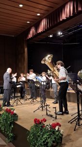 Eine Gruppe junger Blechbläser wird von einem älteren Mann auf einer Bühne dirigiert. Die Musiker spielen Instrumente wie eine große Tuba, Trompeten und eine Posaune und stehen an Notenständern. Die Bühne hat einen Holzboden und einen dunklen Hintergrund. Über ihnen sind rote Vorhänge und eine Holzdecke mit eingelassenen Scheinwerfern zu sehen. Im Vordergrund befinden sich rot-grüne Blumenarrangements.