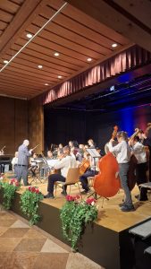Ein Orchester, hauptsächlich bestehend aus jungen Musikern, spielt auf einer erhöhten Bühne in einem Konzertsaal. Ein älterer Dirigent steht mit Taktstock vorne links, dem Orchester zugewandt. Die Musiker spielen verschiedene Instrumente, darunter deutlich erkennbare Celli, Kontrabässe und Violinen; im Hintergrund sind auch Blasinstrumente und Schlagzeug zu sehen. Die Bühne hat einen hellen Holzboden und eine dunkle Rückwand, die von blauen und roten Scheinwerfern beleuchtet wird. Darüber hängt ein dunkelroter Vorhang. Die Decke des Saals ist aus hellem Holz und mit runden Einbauleuchten versehen. Der vordere Rand der Bühne ist mit grünen Hängepflanzen und roten Blumen dekoriert. Der Boden vor der Bühne ist mit quadratischen, gemusterten Fliesen bedeckt.