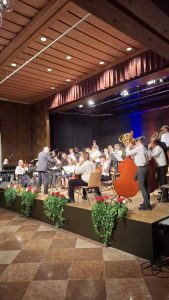 Ein Orchester, bestehend aus überwiegend jungen Musikern in weißen Hemden und einigen Erwachsenen, spielt auf einer Bühne in einem Saal. Ein Dirigent steht im Zentrum der Bühne, mit dem Rücken zum Betrachter, umgeben von Musikern mit Streichinstrumenten (Violinen, Celli, Kontrabässe), Blechblasinstrumenten und einem Klavier. Der Bühnenhintergrund ist dunkel mit einigen farbigen Scheinwerfern und braunen Wandpaneelen. Der Bühnenrand ist mit grünen Pflanzen und roten sowie weißen Blumen geschmückt. Die Decke des Saales ist holzvertäfelt und hat eingelassene runde Lichter sowie eine lange, lineare Leuchtstoffröhre. Der Boden vor der Bühne ist mit glänzenden, rautenförmig verlegten Fliesen in zwei Brauntönen versehen.