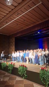 Eine große Gruppe von Kindern und Jugendlichen steht auf einer Bühne und tritt auf, gekleidet in heller Kleidung wie weißen Oberteilen und Jeans. Links sitzt ein erwachsener Mann an einem schwarzen Keyboard mit Mikrofon, der sie dirigiert oder begleitet. Der Bühnenhintergrund ist dunkel und wird von vertikalen blauen und roten Lichtstreifen beleuchtet. Am vorderen Bühnenrand stehen Blumenkästen mit grünen Pflanzen und roten sowie orangefarbenen Blüten. Die Decke ist eine detailreiche Holzpaneeldecke mit eingelassenen Strahlern und einer langen, hängenden Lichtleiste. Der Boden im Vordergrund ist gefliest.