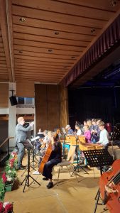 Ein Mann in einem Anzug dirigiert ein Orchester von Kindern und Jugendlichen auf einer Bühne. Im Vordergrund sitzt ein Mädchen und spielt Violoncello. Weitere junge Musiker sind mit verschiedenen Instrumenten wie Xylophonen, Trommeln und Blasinstrumenten (Klarinette) zu sehen, viele an Notenständern. Die Szene findet in einem Saal mit Holzdecke, eingelassenen Lichtern und einem dunklen Bühnenvorhang statt. Links im Vordergrund sind Blumenarrangements und eine Treppe sichtbar.