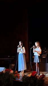 Zwei Frauen stehen mit Mikrofonen auf einer dunklen Bühne, die von Scheinwerfern beleuchtet wird. Die Frau auf der linken Seite trägt ein helles Oberteil und einen langen, plissierten blauen Rock. Die Frau auf der rechten Seite trägt ein helles, schulterfreies Oberteil und einen kurzen, plissierten blauen Rock. Im Hintergrund sind Musikinstrumente und -geräte wie ein Keyboard, Gitarrenverstärker und ein Schlagzeug zu erkennen. Der Vordergrund der Bühne ist mit farbenfrohen Blumenarrangements dekoriert.