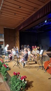 Eine Gruppe von Kindern, ein Jugendorchester, spielt auf einer hölzernen Bühne. Ein älterer Mann in traditioneller Tracht steht links im Vordergrund und leitet die Gruppe. Mehrere Kinder sitzen und spielen Blechblasinstrumente wie Tuben, Trompeten und Posaunen; einige tragen ebenfalls Tracht, darunter Lederhosen und gemusterte Socken. Im Hintergrund sind weitere Instrumente wie ein Schlagzeug, Congas und eine Gitarre zu sehen. Notenständer stehen vor den Musikern. Rechts im Vordergrund lehnt der Korpus eines Kontrabasses. Am vorderen Bühnenrand sind Blumen und Grünpflanzen arrangiert. Der Bühnenhintergrund ist dunkel, mit blauer Beleuchtung rechts. Über der Bühne sind dunkelrote Vorhänge und eine Holzdecke mit runden Einbauleuchten sichtbar.