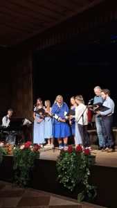 Eine Gruppe von acht Personen tritt auf einer dunklen Bühne auf. Links sitzt ein Mann an einem schwarzen Digitalpiano. Rechts stehen sechs Personen, die meisten halten schwarze Notenmappen und singen in Mikrofone. Eine Frau in einem blauen Kleid und weißem Kopftuch steht prominent im Zentrum. Die vordere Bühnenkante ist mit roten, rosa und orangefarbenen Blumen sowie grünem Efeu und Blattwerk dekoriert. Der Boden vor der Bühne ist mit braun-schwarz karierten Fliesen belegt.