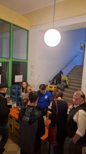 Innenaufnahme einer Veranstaltung oder eines Treffens. Mehrere Personen stehen im Vordergrund um einen Tresen mit Getränken und Flaschen. Links sind grüne Doppeltüren zu sehen. Im Hintergrund führt eine Treppe mit Geländer nach oben. Es ist keine Rampe oder ein Aufzug neben der Treppe sichtbar. Der Boden im direkten Bereich der Menschen ist eben. Getränkekisten stehen teilweise auf dem Boden.