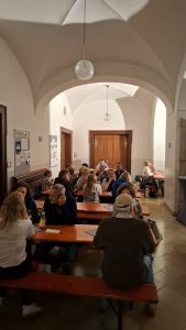 Innenaufnahme eines großen Raumes mit Gewölbedecke, in dem zahlreiche Personen an langen, rotbraunen Holztischen und Bänken sitzen und sich unterhalten. Von der Decke hängen mehrere kugelförmige Glaslampen, die Licht spenden und Schatten werfen. Im Hintergrund sind große Holztüren und an den Wänden Informationsbretter und gewölbte Durchgänge zu sehen. Der Boden ist gefliest.