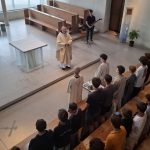 Ansicht von oben in einen hellen, modernen Kirchenraum. Ein Mann in einem goldfarbenen Gewand steht im Altarbereich vor einem gläsernen Altar. Rechts von ihm steht ein junger Mann mit einer E-Gitarre. Eine Gruppe junger Leute, einige davon in hellen Roben, stehen in einer Reihe vor dem Altar. Weitere Personen sitzen auf den Holzbänken im Vordergrund und seitlich. Große, strukturierte Fensterfronten bilden den Hintergrund, und eine Kreuzform ist auf dem Boden zu sehen.