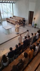Ansicht von oben in einen hellen, modernen Kirchenraum. Ein Mann in einem goldfarbenen Gewand steht im Altarbereich vor einem gläsernen Altar. Rechts von ihm steht ein junger Mann mit einer E-Gitarre. Eine Gruppe junger Leute, einige davon in hellen Roben, stehen in einer Reihe vor dem Altar. Weitere Personen sitzen auf den Holzbänken im Vordergrund und seitlich. Große, strukturierte Fensterfronten bilden den Hintergrund, und eine Kreuzform ist auf dem Boden zu sehen.