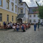Belebter Innenhof unter bewölktem Himmel. Zahlreiche Menschen sitzen an langen Tischen mit roten Platten und Bänken oder stehen in Gruppen. Der Boden ist mit Kopfsteinpflaster ausgelegt. Im Hintergrund sind helle Gebäude mit gelben Akzenten und ein Turm mit Kreuz zu sehen.