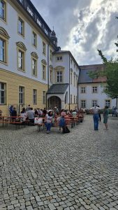 Belebter Innenhof unter bewölktem Himmel. Zahlreiche Menschen sitzen an langen Tischen mit roten Platten und Bänken oder stehen in Gruppen. Der Boden ist mit Kopfsteinpflaster ausgelegt. Im Hintergrund sind helle Gebäude mit gelben Akzenten und ein Turm mit Kreuz zu sehen.