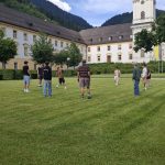 Acht Personen spielen auf einer weitläufigen, grünen Wiese Fußball; ein Ball schwebt in der Luft. Im Hintergrund erstrecken sich große, historische Gebäude mit hellen Fassaden und dunklen Dächern. Rechts ragt ein hoher Kirchturm mit einem grünen Zwiebeldach und einem Kreuz empor. Hinter den Gebäuden erheben sich bewaldete Berge unter einem blauen Himmel mit weißen Wolken.