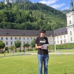 Ein junger Mann steht auf einer großen, grünen Rasenfläche und hält ein gefaltetes Blatt Papier in den Händen. Er trägt ein schwarzes T-Shirt mit Puma-Logo, blaue Jeans und weiße Turnschuhe. Im Hintergrund befindet sich ein langes, helles historisches Gebäude mit vielen Fenstern und einem dunklen Dach, sowie ein hoher Kirchturm mit einer grünen Kuppel auf der rechten Seite. Dahinter erheben sich bewaldete Berge unter einem blauen Himmel mit vereinzelten Wolken. Im Vordergrund steht eine Holzbank mit zwei Getränkeflaschen und eingepackten Gegenständen. Links im Bild ist ein roter Hydrant zu sehen, rechts kleine Fußballtore und Bälle auf dem Rasen.