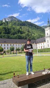 Ein junger Mann steht auf einer großen, grünen Rasenfläche und hält ein gefaltetes Blatt Papier in den Händen. Er trägt ein schwarzes T-Shirt mit Puma-Logo, blaue Jeans und weiße Turnschuhe. Im Hintergrund befindet sich ein langes, helles historisches Gebäude mit vielen Fenstern und einem dunklen Dach, sowie ein hoher Kirchturm mit einer grünen Kuppel auf der rechten Seite. Dahinter erheben sich bewaldete Berge unter einem blauen Himmel mit vereinzelten Wolken. Im Vordergrund steht eine Holzbank mit zwei Getränkeflaschen und eingepackten Gegenständen. Links im Bild ist ein roter Hydrant zu sehen, rechts kleine Fußballtore und Bälle auf dem Rasen.
