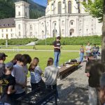 Ein großes, barockes Klostergebäude mit grüner Kuppel und Turm dominiert den Hintergrund unter blauem Himmel. Im Vordergrund versammeln sich etwa 20 Personen auf einer gepflasterten Fläche und Rasen. Ein junger Mann im Zentrum liest aus einem Blatt Papier vor, umgeben von Zuhörern, die auf einer Holzbank sitzen oder stehen. Links im Bild steht ein schwarzer Kohlegrill, um den sich weitere Personen aufhalten.