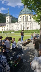 Ein großes, barockes Klostergebäude mit grüner Kuppel und Turm dominiert den Hintergrund unter blauem Himmel. Im Vordergrund versammeln sich etwa 20 Personen auf einer gepflasterten Fläche und Rasen. Ein junger Mann im Zentrum liest aus einem Blatt Papier vor, umgeben von Zuhörern, die auf einer Holzbank sitzen oder stehen. Links im Bild steht ein schwarzer Kohlegrill, um den sich weitere Personen aufhalten.