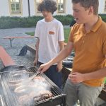 Zwei junge Männer grillen im Freien. Der vordere Mann, in einem orangefarbenen Poloshirt, wendet mit einer Grillzange Würstchen und Burgerpatties auf einem rauchenden Grill. Der andere junge Mann, in einem weißen T-Shirt, steht daneben und schaut zu. Im Hintergrund ist ein großes, helles Gebäude mit vielen Fenstern und grünen Büschen auf einem gepflasterten Hof zu sehen. Unter dem Grill ist ein Sack Grillkohle sichtbar.