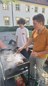 Zwei junge Männer grillen im Freien. Der vordere Mann, in einem orangefarbenen Poloshirt, wendet mit einer Grillzange Würstchen und Burgerpatties auf einem rauchenden Grill. Der andere junge Mann, in einem weißen T-Shirt, steht daneben und schaut zu. Im Hintergrund ist ein großes, helles Gebäude mit vielen Fenstern und grünen Büschen auf einem gepflasterten Hof zu sehen. Unter dem Grill ist ein Sack Grillkohle sichtbar.