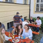 Mehrere junge Menschen sitzen oder stehen an roten Biertischen im Freien. Auf den Tischen stehen Teller mit Essen, Flaschen und Gläser. Eine junge Frau im linken Vordergrund blickt in die Kamera. Im Hintergrund ist ein helles Gebäude mit gelben Fensterrahmen und Gitterstäben an den Fenstern zu sehen. Der Boden ist mit Kopfsteinpflaster ausgelegt.