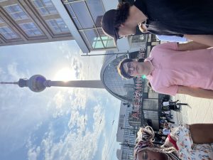 Alexanderplatz unter einem blauen Himmel mit vereinzelten Wolken. Die Sonne strahlt hell hinter dem Turm hervor. Links und rechts sind weitere Personen teilweise im Bild.