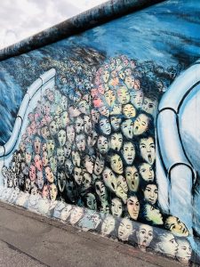 dichte Ansammlung stilisierter menschlicher Gesichter darstellt. Die Gesichter haben verschiedene Hauttöne (blass, gelblich, rötlich, bläulich) und zeigen unterschiedliche Ausdrücke, einige mit offenen Mündern, andere mit geschlossenen Augen. Sie scheinen aus oder durch einen fließenden, wirbelnden blauen Hintergrund aufzusteigen. Zwei große, helle, rohr- oder armähnliche Formen umrahmen die Gesichtermassen von links und rechts. Am unteren Bildrand ist ein graubrauner, asphaltierter Boden zu sehen, der leicht nach links ansteigt.