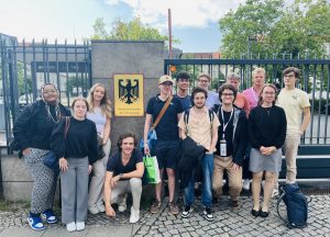 Eine Gruppe von überwiegend jungen Erwachsenen steht und hockt vor einem hohen schwarzen Metallzaun und einer Mauer. An der Mauer ist ein großes gelbes Schild mit dem Bundesadler und der Aufschrift "Bundesministerium der Verteidigung" angebracht. Im Hintergrund sind Bäume und weitere Gebäude zu sehen. Der Boden ist mit Kopfsteinpflaster ausgelegt. Die meisten Personen lächeln in die Kamera.