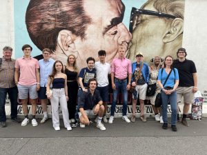 Eine Gruppe von 15 Personen, darunter Männer und Frauen unterschiedlichen Alters, steht lächelnd vor einem großen Wandgemälde. Das Gemälde zeigt die Köpfe zweier Männer, die sich küssen, vor einem hellblauen Hintergrund. Die Personen tragen Freizeitkleidung, ein Mann hockt in der Mitte im Vordergrund. Links und rechts des Gemäldes ist eine weiße Mauer sichtbar. Der Boden ist ein grauer, asphaltierter Weg mit Rissen.