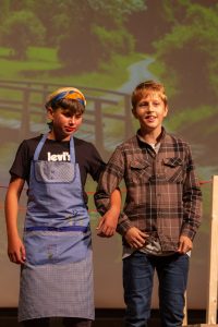 Zwei Jungen stehen Arm in Arm auf einer Bühne. Der linke Junge trägt eine blaue Schürze, ein gemustertes Kopftuch und roten Lippenstift. Der rechte Junge trägt ein braun kariertes Hemd und Jeans. Beide schauen in die Kamera. Im Hintergrund ist eine unscharfe Projektion einer grünen, baumbestandenen Landschaft mit einer Straße und einer Brücke zu sehen.